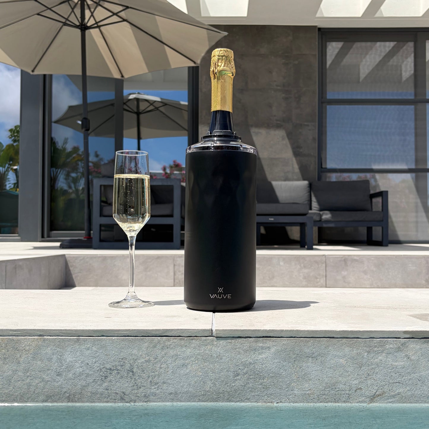 Bottle of champagne in matte black insulated wine chiller at the poolside, with a modern villa in the background.